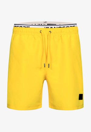 Muskelbepackter Mann barfuß, lächelnd und nach unten schauend, trägt leuchtend gelbe Badeshorts mit schwarzem Kordelzug auf schlichtem hellem Hintergrund.