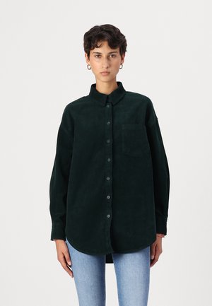 Tummanvihreä corduroy-paita, jossa on napitus etuosa, viistot kaulus ja rintatasku. Siinä on pitkät hihat ja pyöreä helma.