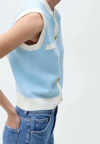 Chaleco de punto azul claro con bordes acanalados blancos y botones dorados. Presenta un diseño ajustado y sisas abiertas, combinado con jeans azules.