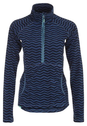 Kvinde iført en blå fleece med zigzag-mønster og halv lynlås samt sorte udendørsbukser, stående med den ene hånd i lommen.