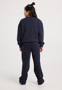 Mujer joven con cabello oscuro en un semi-recogido, vistiendo un sudadera azul marino y pantalones de chándal a juego, de pie de espaldas sobre un fondo liso.