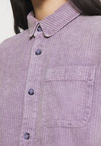 Lila kordflöjtskjorta med krage, framknappar och ficka med präglad logotyp. Strukturerad ribbad textil med släta knappar.