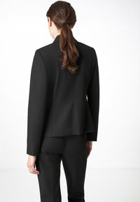 Černé sako na míru s mírně rozšířeným zadním dílem, dlouhými rukávy a minimalistickým designem. Vyrobeno z hladké látky, bez viditelných kovových prvků nebo vzorů.