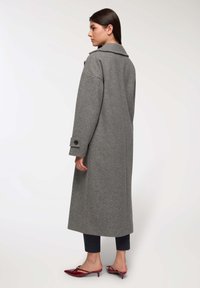 Kobieta stojąca w profilu, ubrana w długi szary płaszcz, ciemne, krótkie spodnie i czerwone buty na niskim obcasie w białym tle.