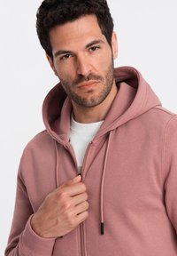 Sweat à capuche zippé en rose poussiéreux, en mélange de coton. Comprend des cordons de serrage ajustables et des poignets côtelés, avec un col rond blanc en dessous.