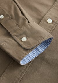 Mâneca unei cămăși din bumbac maro, cu butoane albe și căptușeală cu dungi albastre. Textură netedă, design standard și cusături lucrate cu atenție.