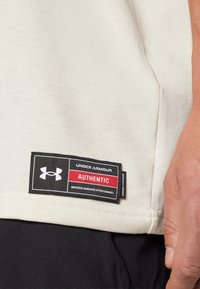 Ljusbeige bomullströja med en svart och röd etikett som säger "Under Armour Authentic" sydd nära fållen, som visar en mjuk textur.