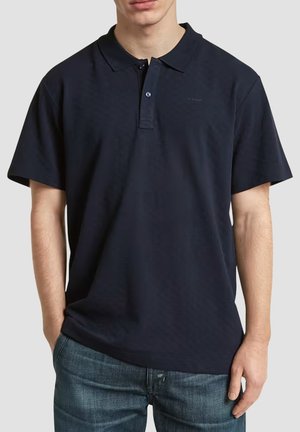 Homme portant un polo bleu marine uni à manches courtes et un jean bleu, debout avec une main dans la poche.