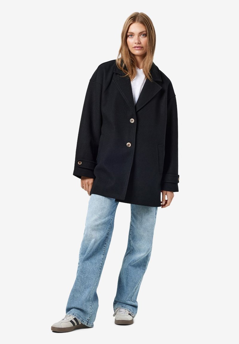 Schwarzer Wollmix-Übermantel im Oversized-Stil mit großen Revers, zwei Fronttaschen und Knopfverschluss. Kombiniert mit hellen Jeans und Turnschuhen.