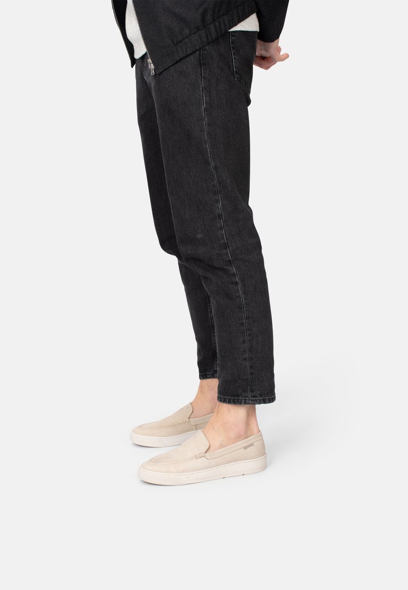 Scarpe slip-on beige con una texture in suede e dettagli di cucitura discreti, abbinate a jeans neri a gamba dritta. Primo piano sulle calzature.