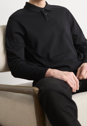Personne portant un polo à manches longues noir et un pantalon noir, assise sur une chaise beige rembourrée avec des accoudoirs en bois.