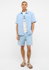 Camisa de mezclilla azul claro de manga corta con un bolsillo en el pecho, combinada con pantalones cortos a juego. El modelo lleva sandalias marrones. Diseño simple y casual.