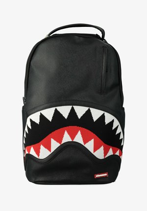 Mochila negra con un diseño de dientes de tiburón en rojo, blanco y negro en la parte frontal y una pequeña etiqueta del logo "Sprayground" en rojo cerca de la parte inferior.