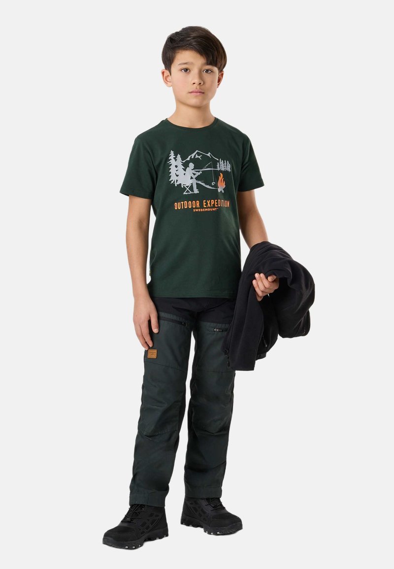 Αγόρι που φορά σκοτεινό πράσινο T-shirt "Outdoor Expedition" και παντελόνι πεζοπορίας, κρατώντας μια μαύρη ζακέτα, στέκεται μπροστά σε λευκό φόντο.