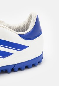 Scarpa sportiva bianca con accenti blu, dotata di un tallone testurizzato, una suola prominente con tacchetti per la trazione e linee di design curve.