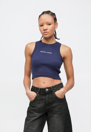 Ungdom med flettet hår iført en marineblå kort topp med "Red Bull Racing" og svarte jeans, hendene i lommene, nøytralt uttrykk.