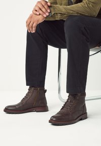 Bottines marron en cuir avec bouts ronds, lacets à l'avant et fermeture éclair latérale. Accent marron texturé au niveau du revers. Semelle en caoutchouc avec crampons.