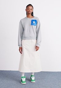 Szürke pulóver kék logós zsebbel, krémszínű maxi szoknya, fehér zoknik, zöld sportcipők. Puha anyag, laza szabás. Minimalista design.