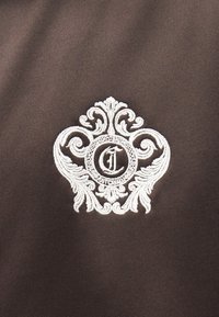 Λεπτομερές άσπρο κεντημένο έμβλημα με σχέδια τύπου καμπυλών και το γράμμα "J" μέσα σε κυκλικό πλαίσιο πάνω σε σκουρό καφέ ύφασμα.