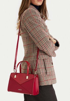 Vrouw draagt een beige geruite blazer met rode lijnen en heeft een rode leren handtas met handvatten bovenop en een schouderriem aan haar zijde.