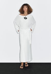 Weißes Leinenkleid mit weitem Ausschnitt und langen Ärmeln, die in der Taille gebunden sind. Komplettiert wird das Outfit durch eine schwarze Anhänger-Halskette und flache braune Sandalen.