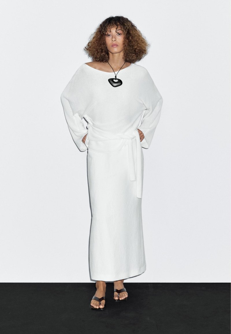 Weißes Leinenkleid mit weitem Ausschnitt und langen Ärmeln, die in der Taille gebunden sind. Komplettiert wird das Outfit durch eine schwarze Anhänger-Halskette und flache braune Sandalen.