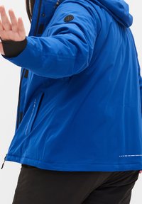 Chaqueta aislante azul con capucha, acentos negros, un logo circular en la manga y detalles de costura visibles en el dobladillo y bolsillos.