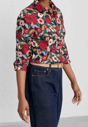 Kobieta ubrana w czerwoną, różową i zieloną koszulę w kwiaty na guziki z podwiniętymi rękawami, włożoną do wysokich, ciemnoniebieskich dżinsów z plecionym beżowym paskiem.