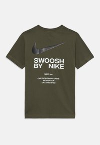 Alyvuogių žalios spalvos trumpomis rankovėmis marškinėliai, pagaminti iš medvilnės. Puošia juodas Nike Swoosh logotipas ir baltas tekstas ant nugaros, apačioje – priežiūros informacija.