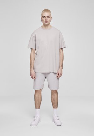 Mladi moški stoji obrnjeno proti gledalcu, oblečen v svetlo sivo preveliko majico, ujemajoče kratke hlače, bele nogavice in bele superge na enobarvnem ozadju.