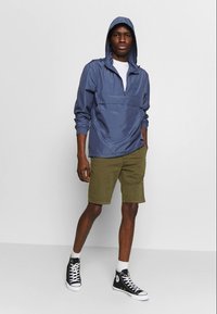 Chaqueta con capucha de color azul claro y cremallera en el frente, combinada con pantalones cortos de color verde oliva y zapatillas altas negras.