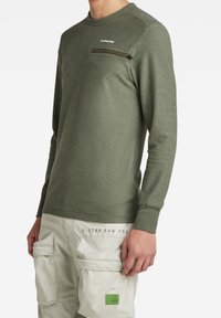 Olivgrön långärmad skjorta med en dragkedja på bröstfickan, ribbad halsringning och åtsittande ärmslut, kombinerad med beiga cargobyxor med fickor.