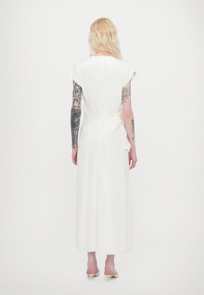 Weißes, knöchellanges Kleid mit kurzen, angeschnittenen Ärmeln, drapierten Details an der Taille und glatter Stoffstruktur. Getragen mit spitzen, beigen Absätzen.