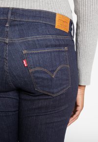 Primer plano de una persona que lleva vaqueros Levi's azul oscuro con una etiqueta roja y un parche de cuero naranja de la marca, combinados con un suéter de canalé gris claro.