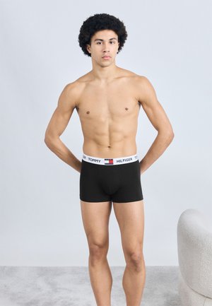 Mustad puuvillased bokserid valge vööümbrisega, millel on logo "TOMMY HILFIGER." Modell seisab neutraalse värvitooniga taustal.