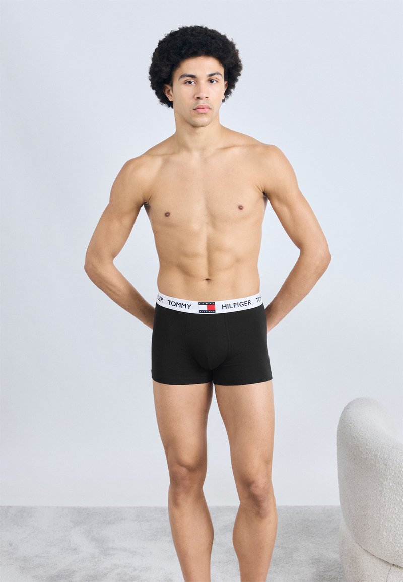 Μαύρα βαμβακερά μπόξερ με λευκή ελαστική μέση που φέρει το λογότυπο "TOMMY HILFIGER". Το μοντέλο στέκεται μπροστά σε ουδέτερο φόντο.