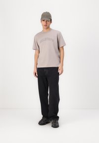 Γκρι κοντομάνικο μπλουζάκι με το κείμενο "BELSTAFF", μαύρα φαρδιά τζιν παντελόνια και γκρι καπέλο με το κείμενο "Won". Μαύρα αθλητικά παπούτσια με σχέδιο.