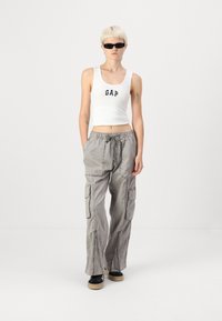 Débardeur blanc court avec le logo "GAP" bleu ; pantalon cargo gris avec plusieurs poches et zips ; chaussures noires avec une semelle beige.