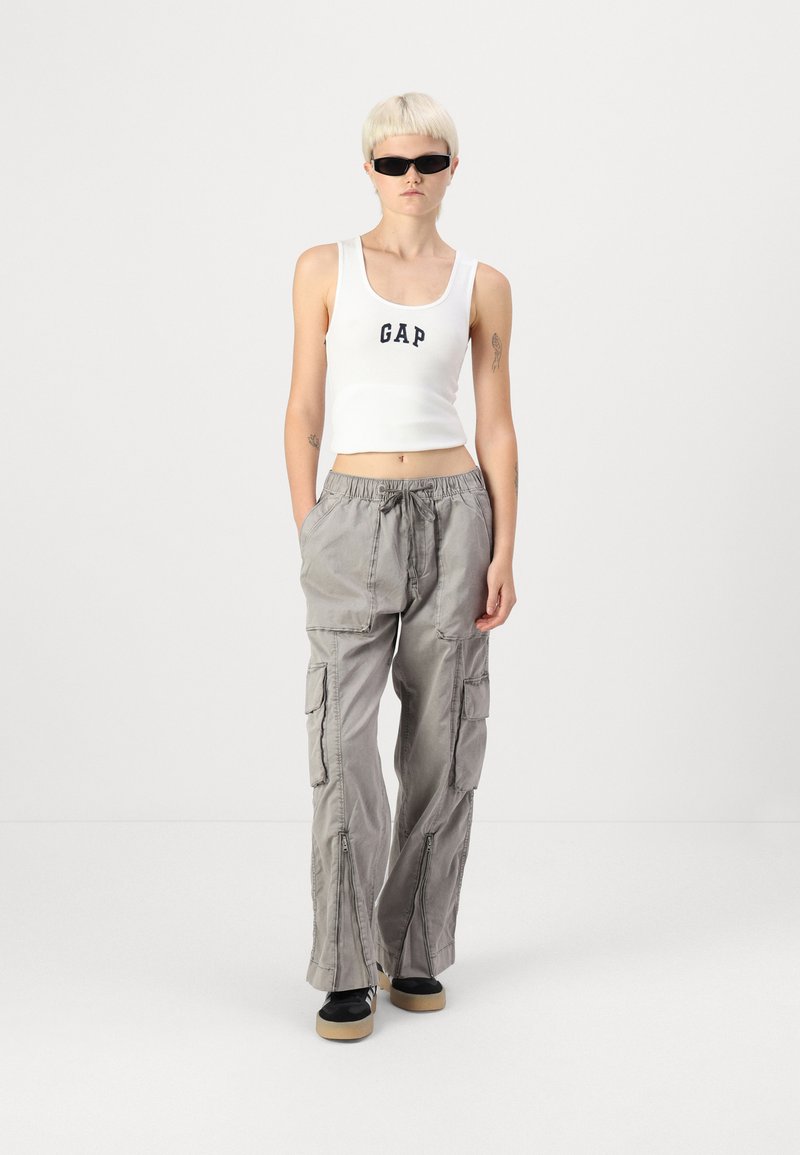 Top corto blanco con el logo de "GAP" en azul; pantalones cargo grises con múltiples bolsillos y cremalleras; zapatos negros con suela beige.