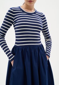 Haut à manches longues rayé bleu marine et blanc, associé à une jupe unie bleu marine. La robe présente une taille froncée et des poches latérales.