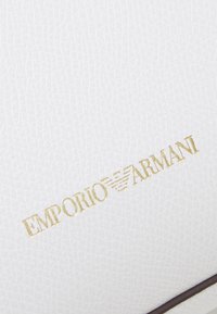 White textured surface with a gold "Emporio Armani" logo and a small emblem. Accented with a dark brown trim along the edge.