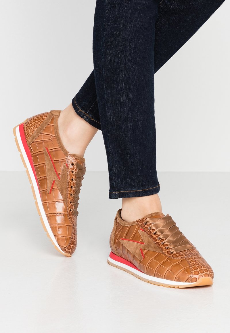 Bruna sneakers med krokodilstruktur och röda accenter, med snörningsdesign och en vit gummisula, paret med mörka jeans.
