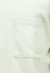 Κοντινή λήψη από τσέπη από ανοιχτό μπεζ ύφασμα με κεντημένο λογότυπο λουλουδιού και το κείμενο "HUGO" σε ένα ένδυμα.