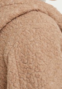 Tela marrón con textura y un acabado suave y esponjoso. La prenda presenta un diseño en capas, destacando su calidez y cualidad táctil.