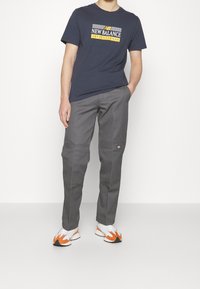 Homme portant un t-shirt New Balance bleu marine, un pantalon gris avec des détails au niveau des genoux, et des baskets blanches avec des accents orange, debout devant un fond blanc.