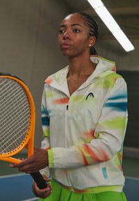 White zip-up jacket with colorful abstract patterns in green, orange, and blue, paired with a green skirt; holding an orange tennis racket.