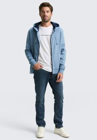 Lichtblauwe zip-up hoodie met een voorzak, gedragen over een wit T-shirt met zwarte tekst, gecombineerd met een donkere spijkerbroek en witte sneakers.