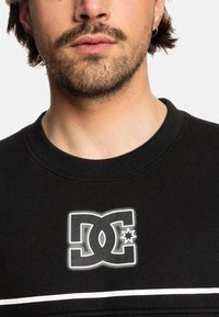 Black sweatshirt featuring a prominent white DC logo on the chest, with a round neckline and horizontal white stripe across the front.