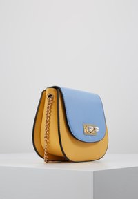 Bolso cruzado de cuero azul claro y amarillo con un cierre de herrajes en tono dorado, correa de cadena y forma redondeada que presenta texturas suaves.
