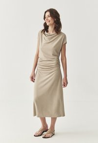 Abito beige con scollo a cowl e maniche corte, dettaglio arricciato in vita e gonna fluida. Indossato con sandali di colore chiaro. Tessuto con una texture liscia.